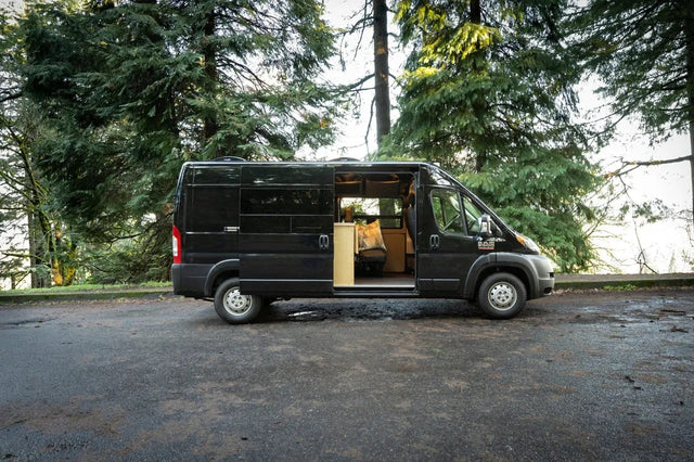 Black Ram ProMaster camper van RV rental parked among tall pine trees with side door open, showing the converted interior.