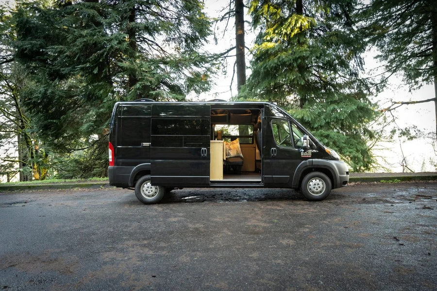 Black Ram ProMaster camper van RV rental parked among tall pine trees with side door open, showing the converted interior.