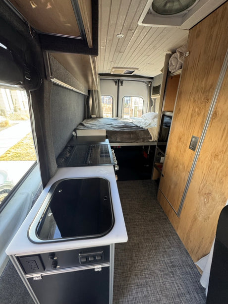 RV rental interior: raised bed, compact kitchen with sink/stove, ample storage, and a textured floor.