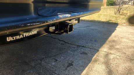 Close-up of a vehicle's rear bumper and Ultra Frame trailer hitch receiver, ideal for an RV rental. Concrete driveway visible.