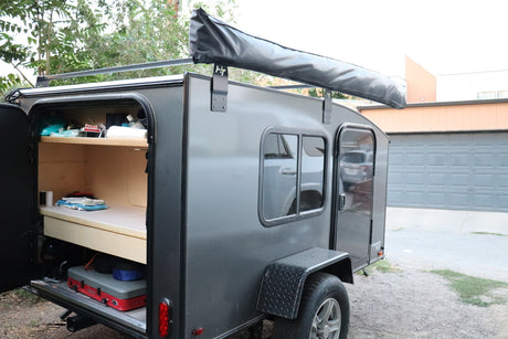 This compact gray RV rental features an open rear hatch revealing a built-in bed, shelving, & camping stove.