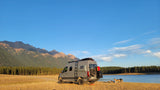 Grey RV rental parked on golden grass by a blue lake with dogs. Majestic mountains in the distance under clear skies.