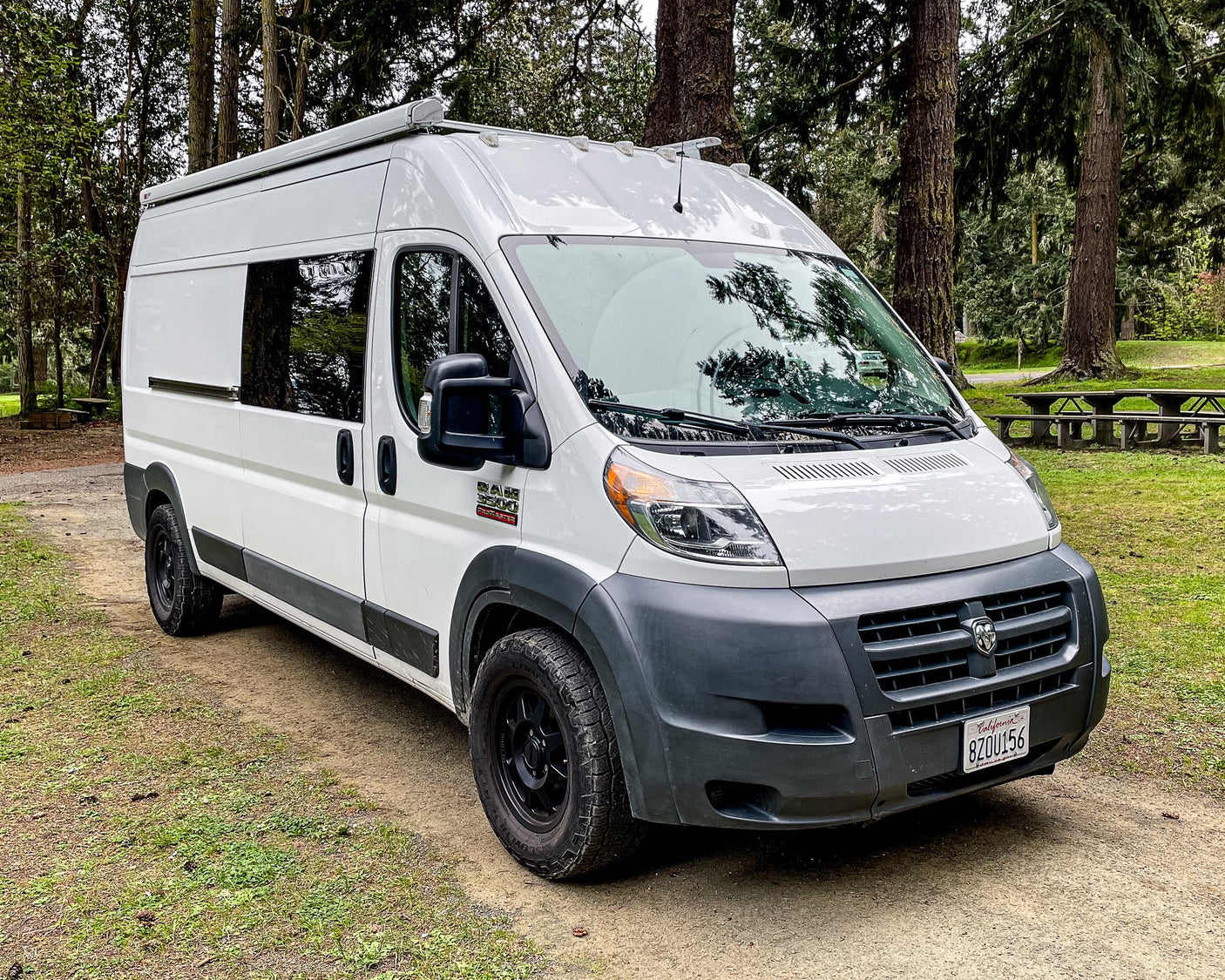 White Ram ProMaster camper van RV rental parked in a forest, ideal for van life. Picnic tables visible. CA plate 8ZOU156.