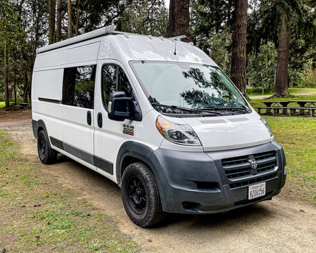 White Ram ProMaster camper van RV rental parked in a forest, ideal for van life. Picnic tables visible. CA plate 8ZOU156.