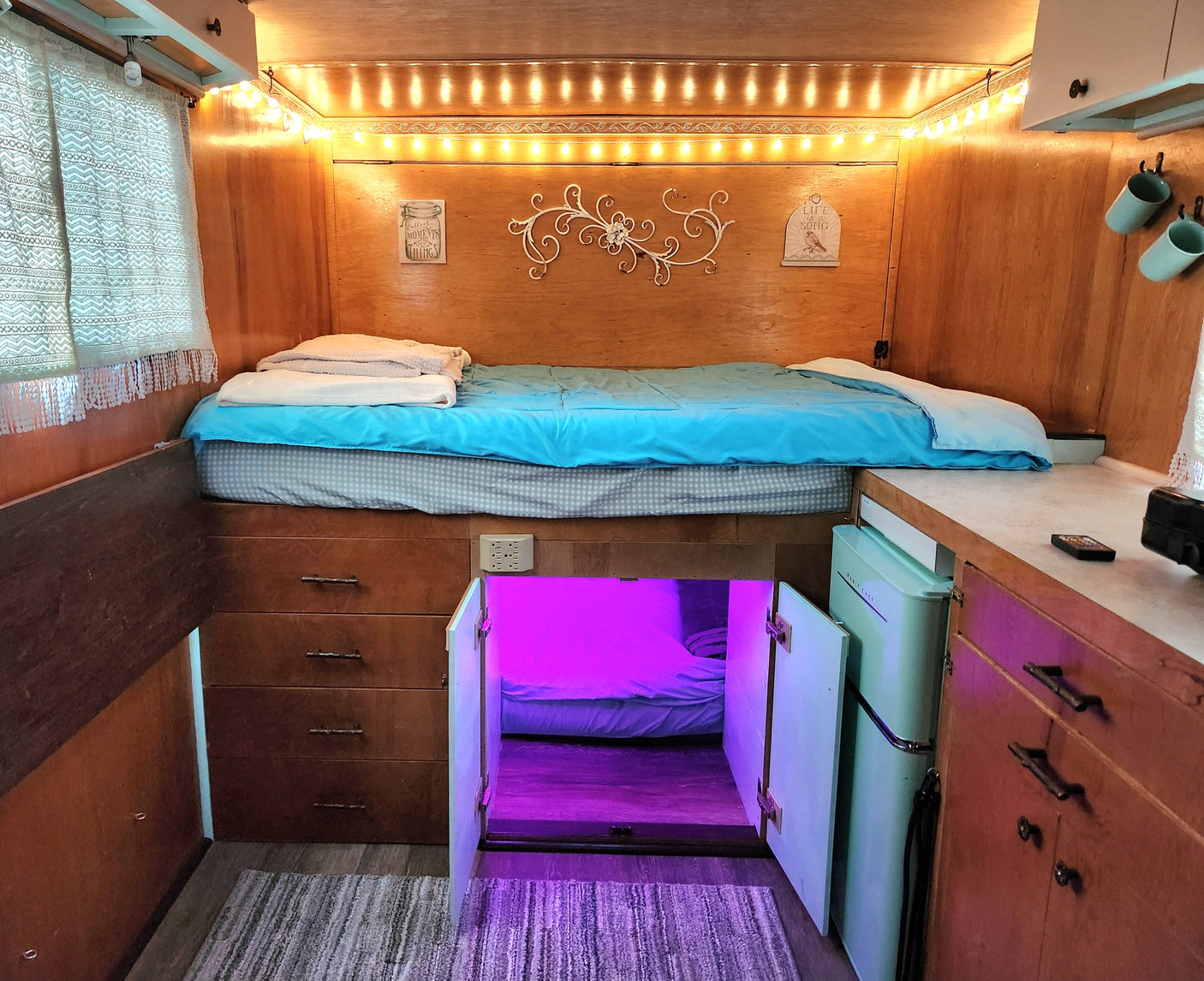 Cozy RV rental interior with a raised bed over storage, compact kitchen counter, and a unique hidden lower bunk with purple lighting.