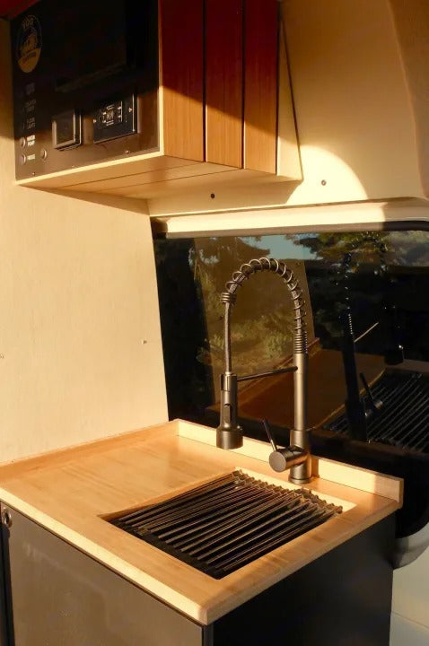 Inside an RV rental, a bright kitchen featuring wood counters, a modern faucet, roll-up sink drainer, and a control panel for water/lights.
