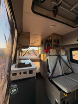 Spacious RV rental interior with kitchen, bed, and passenger seats, offering desert views through the open rear doors.