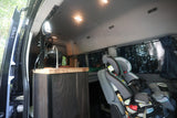 Family-ready RV rental interior: shows a compact kitchen counter, fan, and two child car seats.