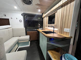 Modern RV rental conversion: L-shaped sofa, full kitchen with sink/cooktop, ambient lighting. Ideal for road trips.
