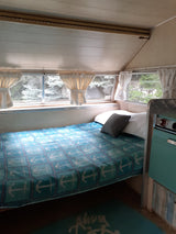 This cozy rv rental interior features a bed with an anchor blanket, bright windows showing trees, and a teal kitchen unit.