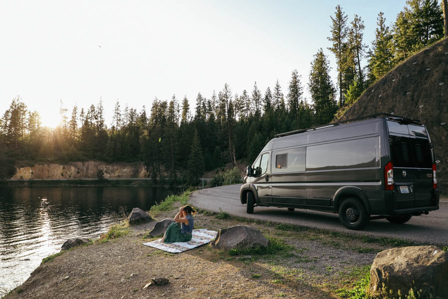 Enjoy nature with an RV rental. A dark gray camper van sits by a lake with a person relaxing on a blanket at sunset.