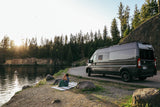 Enjoy nature with an RV rental. A dark gray camper van sits by a lake with a person relaxing on a blanket at sunset.