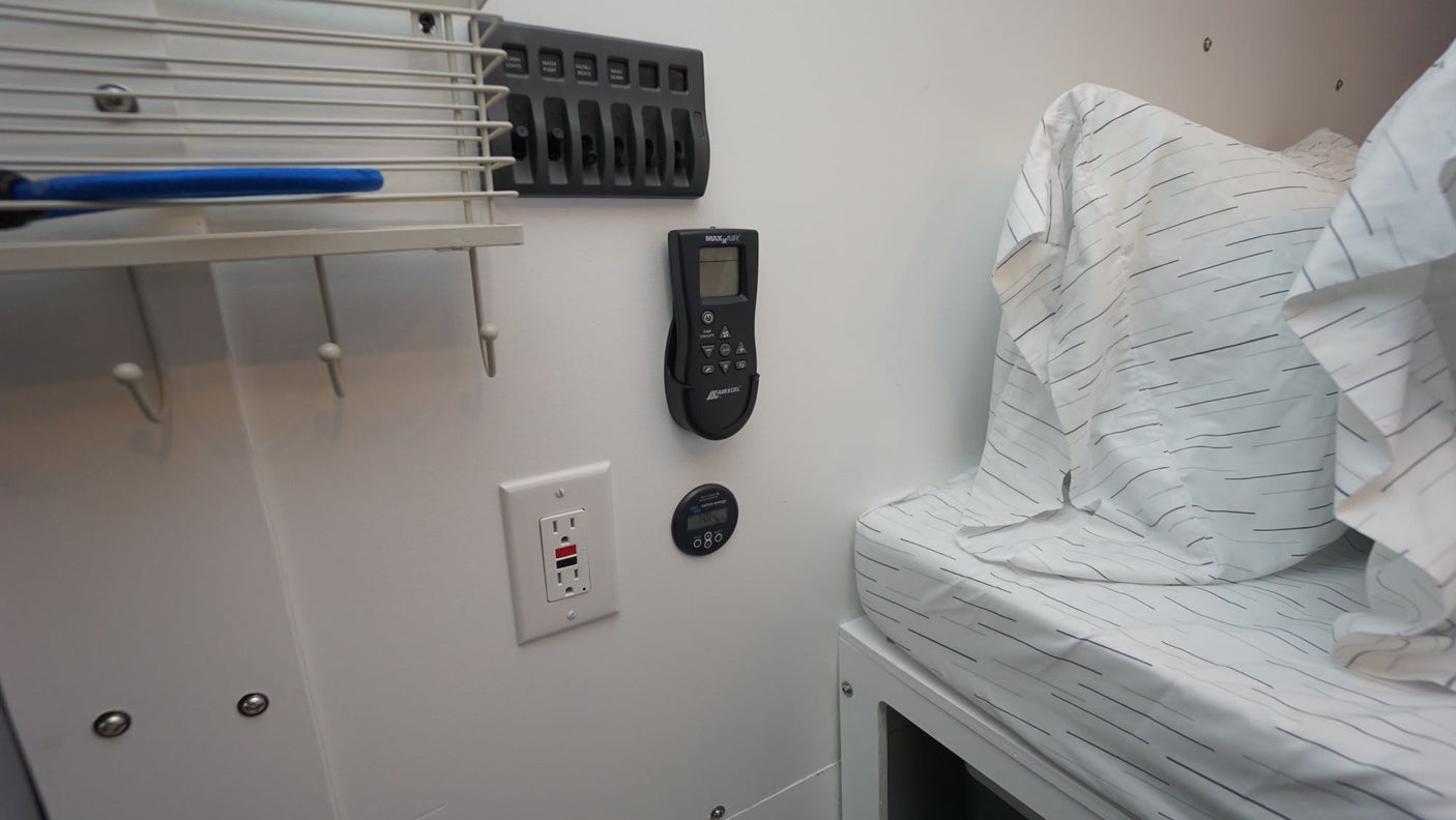 Inside an RV rental: Bed with striped sheets, wall-mounted AC remote, power panel, and storage hooks offer convenience.