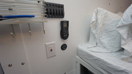 Inside an RV rental: Bed with striped sheets, wall-mounted AC remote, power panel, and storage hooks offer convenience.