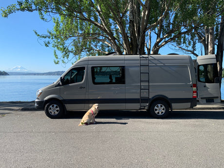 Scenic view of a grey RV rental van parked by water with a dog, Mt. Rainier visible, under a clear sky.