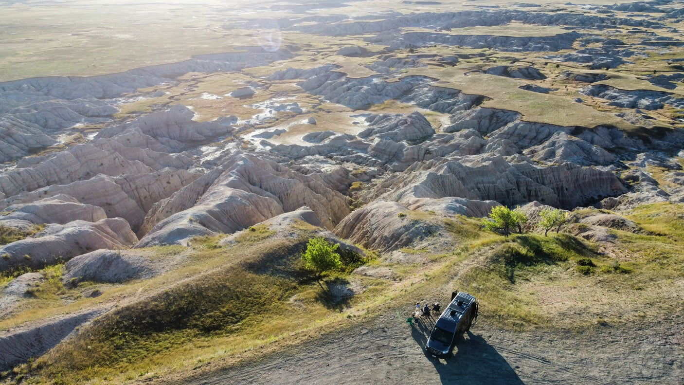 An RV rental parked at a scenic overlook, offering a high-angle view of vast, eroded badlands with people enjoying the stunning landscape.