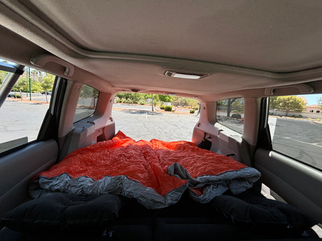 A car's interior transformed into a cozy sleeping space for camping, complete with a bright orange sleeping bag. Your RV rental alternative!