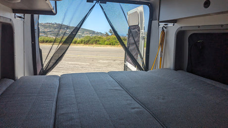Inside a spacious RV rental, a comfortable bed faces open rear doors with a mesh screen & scenic outdoor view.