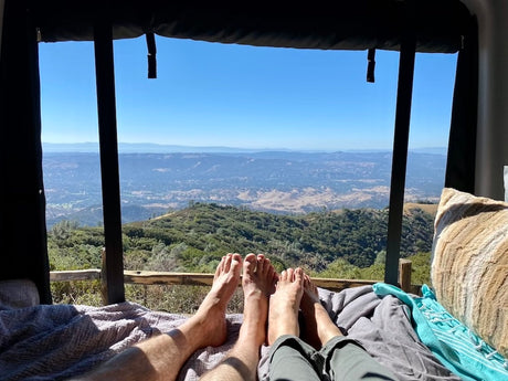Couple's feet up, enjoying an expansive mountain and valley view from a cozy RV rental bed. Perfect escape.