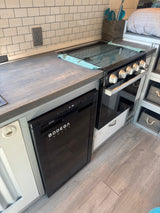 Compact RV rental kitchen with a cooktop/oven, cooler, wood-look countertop, and subway tile backsplash. Ready for travel!