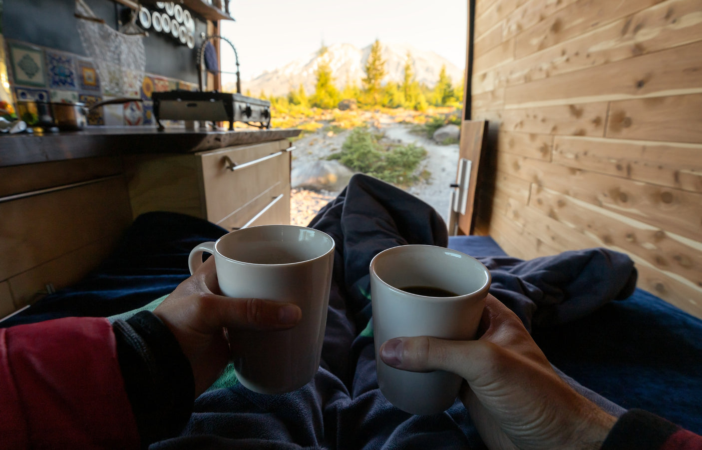 Cozy morning in an RV rental: hands hold coffee, facing open doors to a sunlit mountain view and kitchen area.