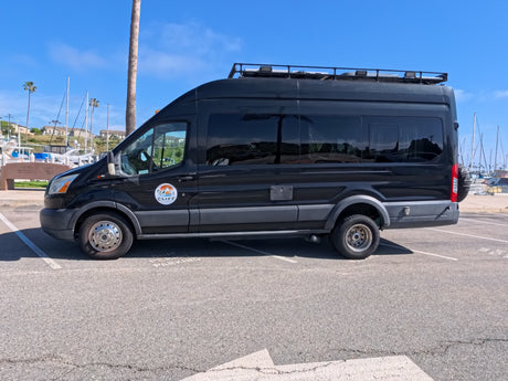 Stylish black custom camper van, likely an rv rental, parked by a sunny marina with boats and palm trees.