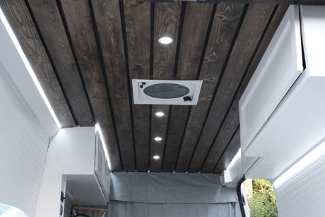 RV rental interior: Dark wood plank ceiling, bright recessed lights, a central fan, and white overhead cabinets with strip lighting.