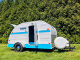 A charming white & blue Retro Jr. compact teardrop travel trailer for rv rental sits on grass, door open to blue interior.