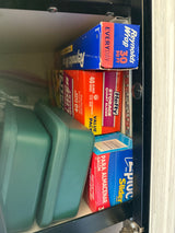 RV rental kitchen cabinet with organized food storage: Reynolds Wrap, Hefty, Ziploc bags, and green containers.