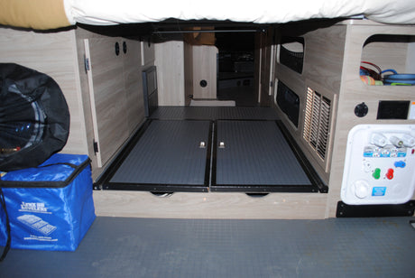 Inside an RV rental: organized under-bed storage with textured floor, access hatches, and a visible water utility panel.