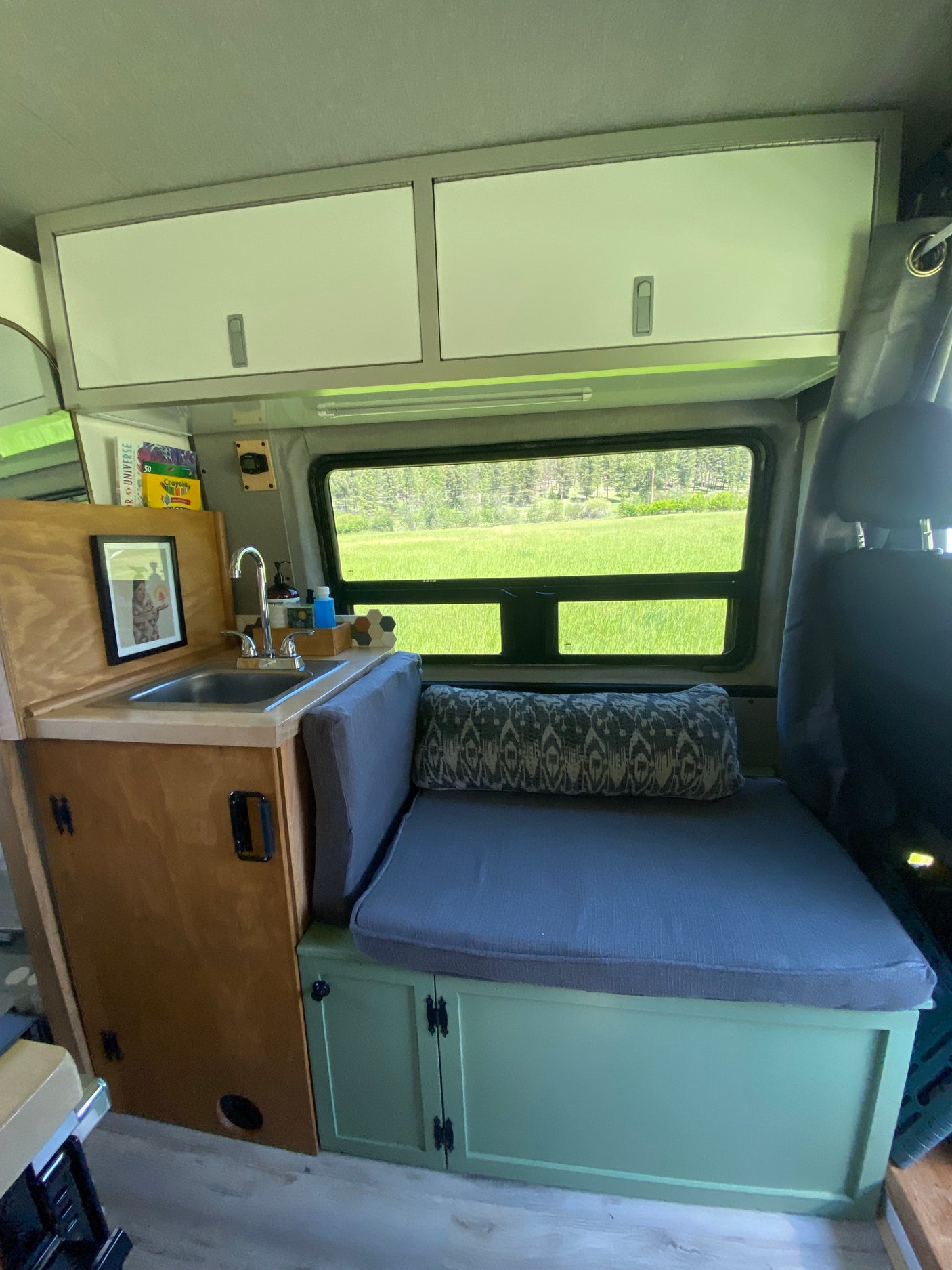 RV rental camper van interior. Includes a compact kitchenette, sink, overhead storage cabinets, and a grey bench seat with built-in storage.