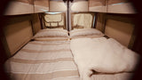 Inside an RV rental: a neat, comfortable bed with striped bedding & a fluffy white blanket, ready for travel.