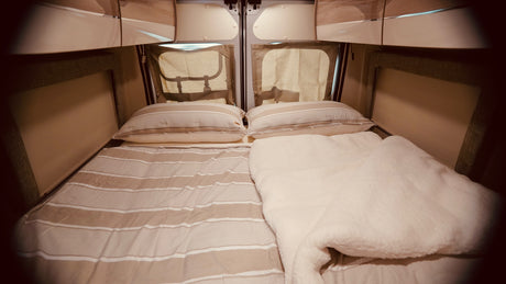 Inside an RV rental: a neat, comfortable bed with striped bedding & a fluffy white blanket, ready for travel.