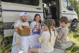 Smiling family enjoys an outdoor picnic by their RV rental camper van, with food & drinks. Perfect for family adventures!