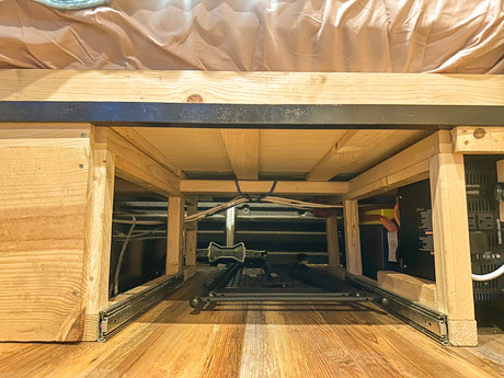 Under-bed RV rental storage with battery, inverter, and pull-out tray setup. Wood frame and electrical system visible.