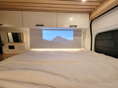 Relax in your cozy rv rental with a projector screen displaying mountains, overhead cabinets, and a comfortable bed.