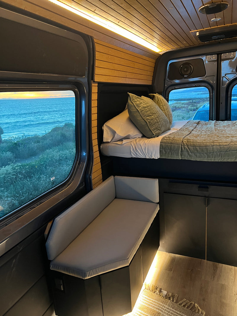 rv rental: Interior of a cozy campervan with a bed, bench, wood ceiling, and stunning ocean view at sunset. — photo 13