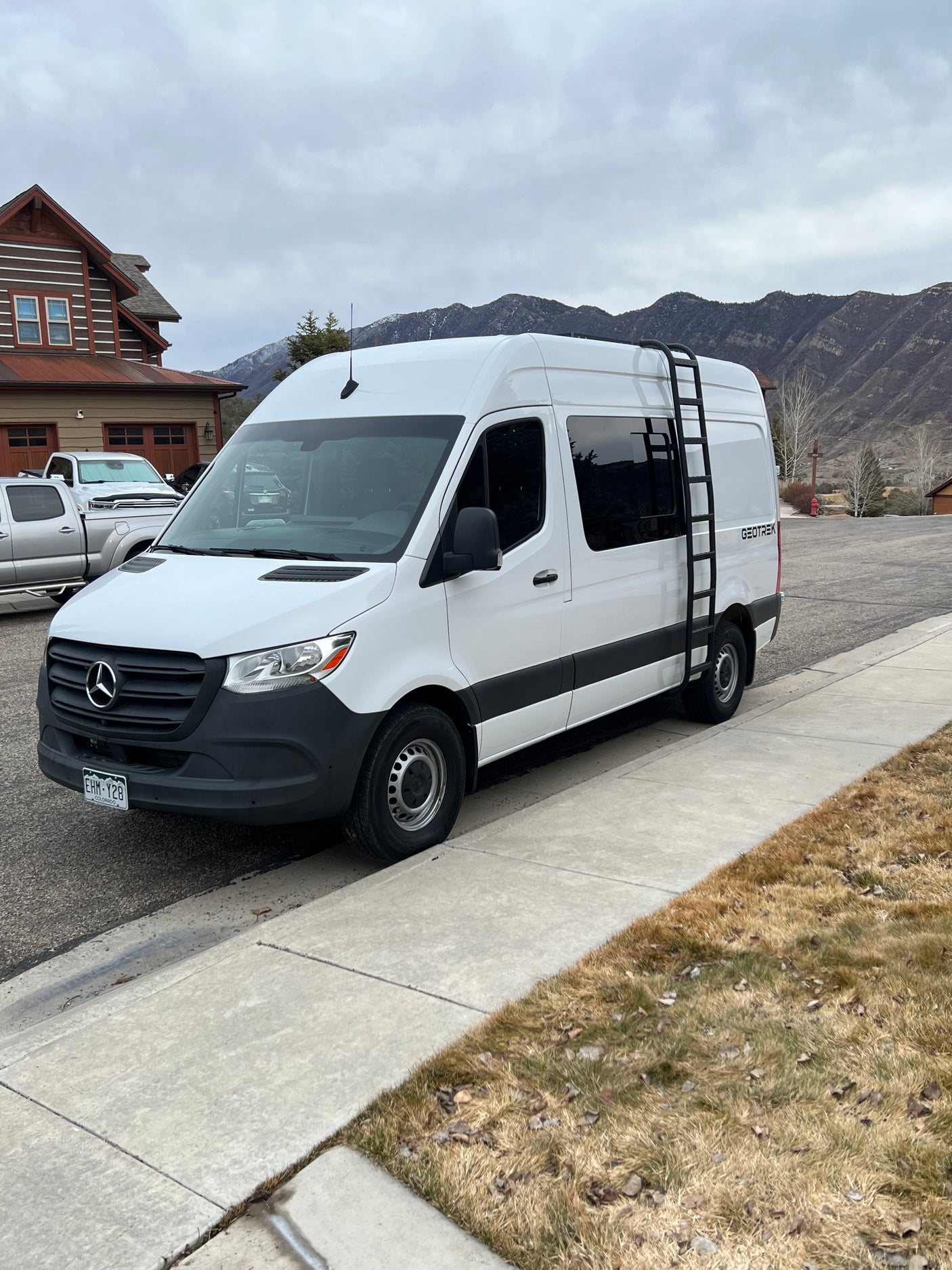 White Mercedes-Benz Sprinter van by GEOTREK, likely an RV rental, parked roadside with mountains in view.