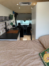 Cozy RV rental interior featuring a gingham bed, modern kitchen with a sink & backsplash, and a view into the front cabin.