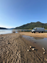  parked on a beach by a lake and mountains. Great RV rental for adventure.|For RV rental: This AWD 170 camper van offers a bed