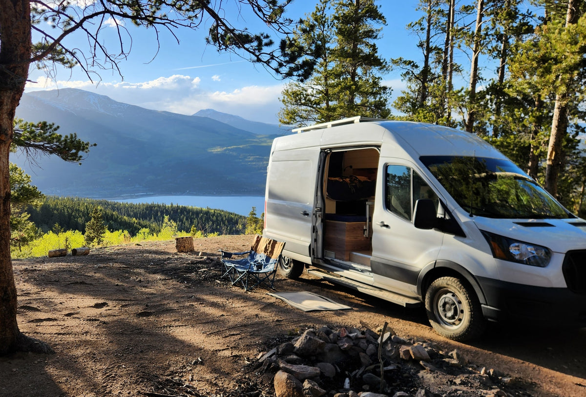 RV rental adventure: White camper van parked on a scenic mountain overlook with a lake view. Chairs and fire pit ready.