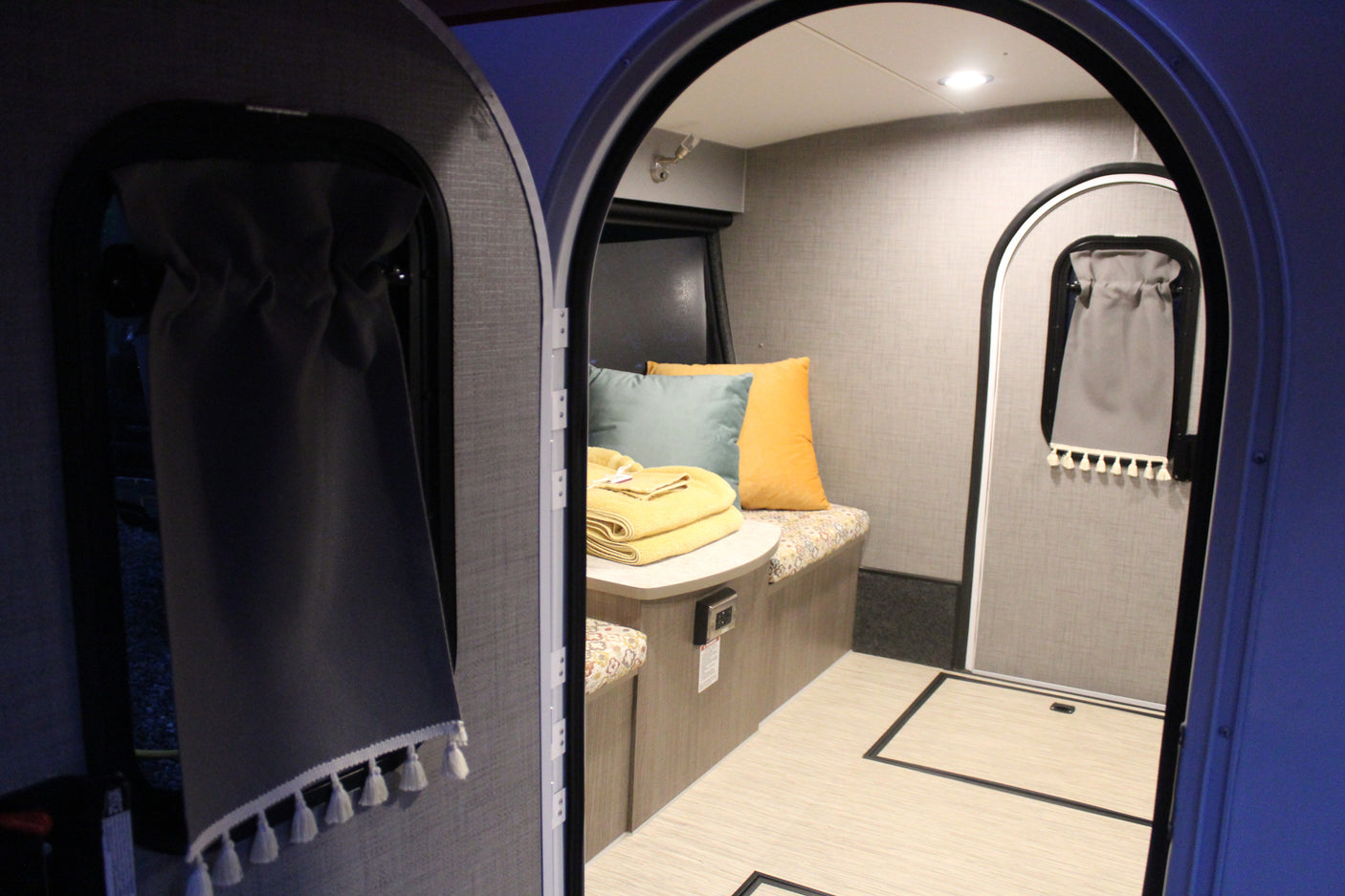 Inside a modern RV rental, a compact living space is visible through arched doorways, featuring a bench, table, and soft furnishings.