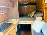 Cozy wood-paneled RV rental interior with a comfy bed, modern kitchen cooktop, and microwave. Ideal for your next adventure.