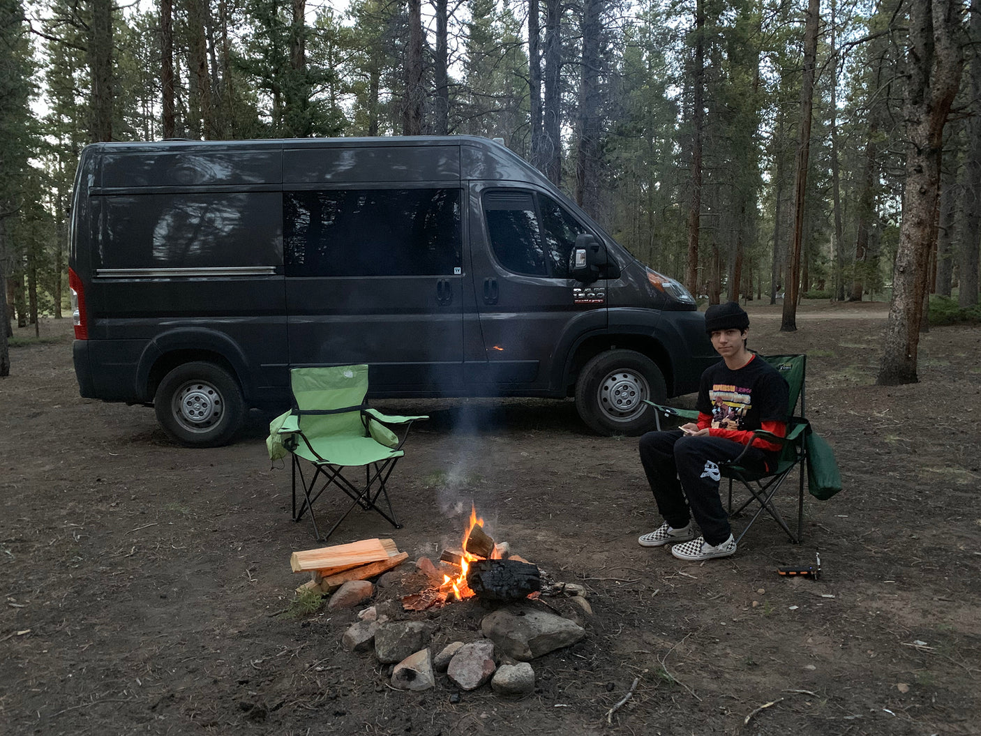RV rental camper van parked in a forest campsite. A person sits by a crackling campfire, enjoying the outdoors.