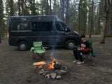 RV rental camper van parked in a forest campsite. A person sits by a crackling campfire, enjoying the outdoors.