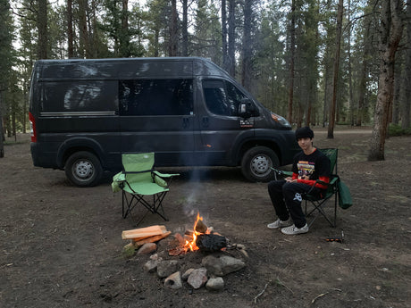 RV rental camper van parked in a forest campsite. A person sits by a crackling campfire, enjoying the outdoors.