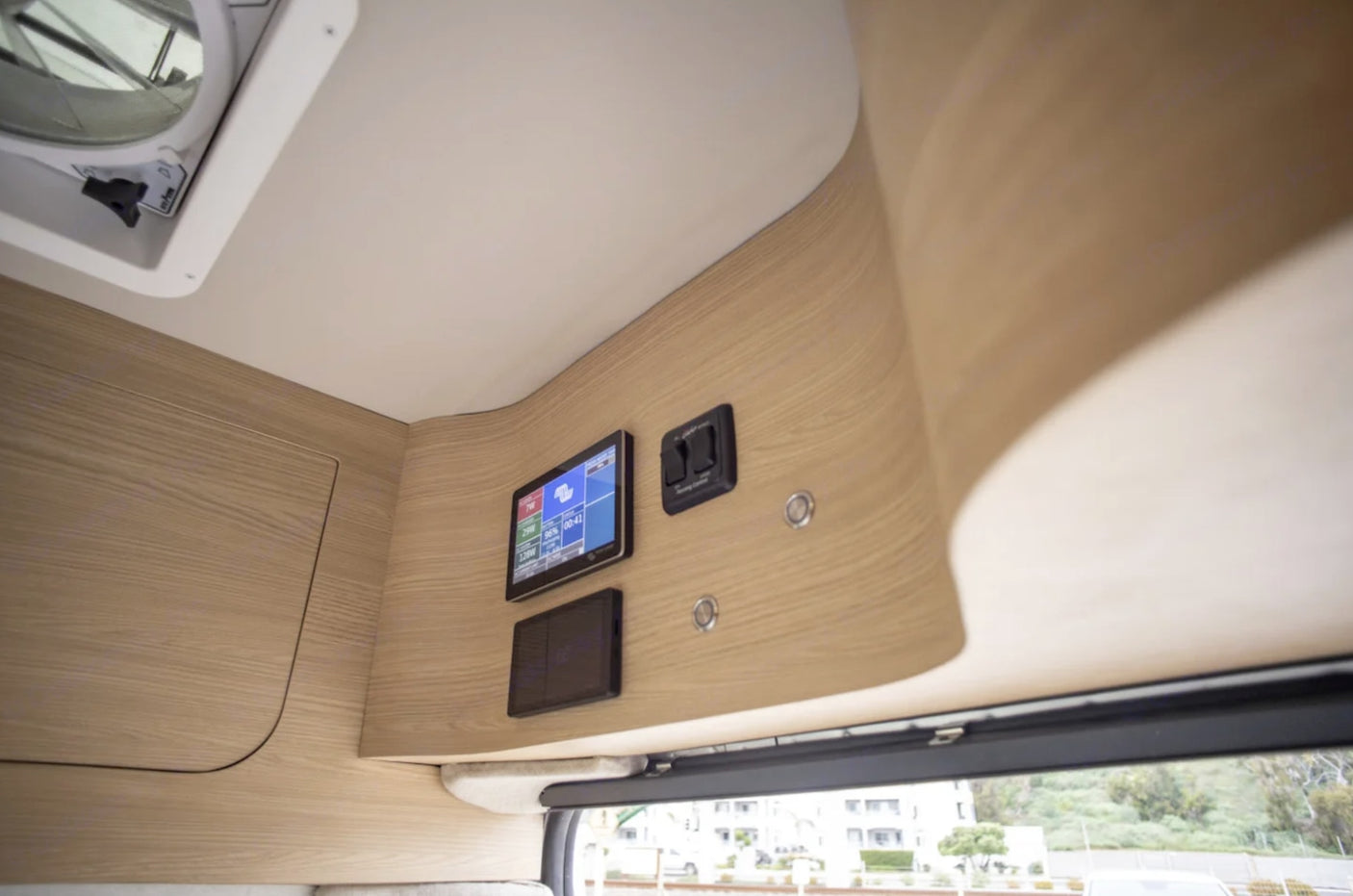 RV rental interior: light wood paneling, digital control screen, and lighting switches visible. A ceiling vent is also present.