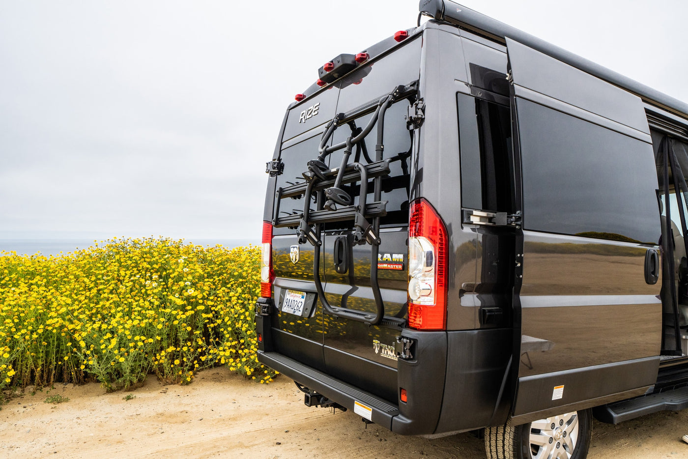 Dark grey Thor Rize RV rental, a Ram ProMaster camper van with a bike rack, parked among yellow wildflowers by the ocean. — photo 5