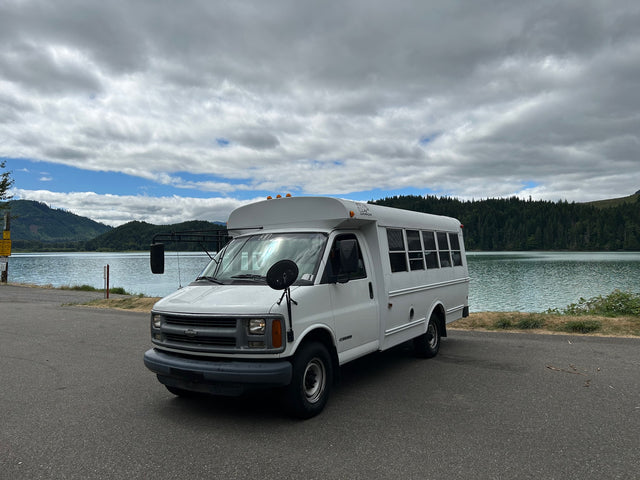 White short bus RV rental by a serene lake and forested mountains under cloudy skies. Perfect for scenic road trips.
