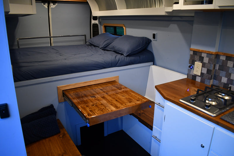 Compact RV rental interior with a comfy bed, pull-out table, and full kitchenette including stovetop and tile backsplash. — photo 7